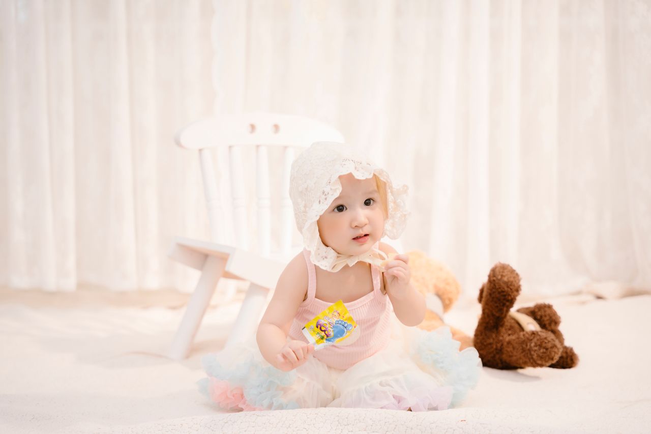 gallery-4 Adorable baby girl in a bonnet enjoying a lollipop with a teddy bear, set in a soft indoor setting.