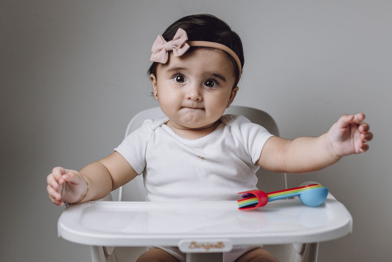 services-02 Cute baby girl in high chair with colorful toy utensils against gray background.
