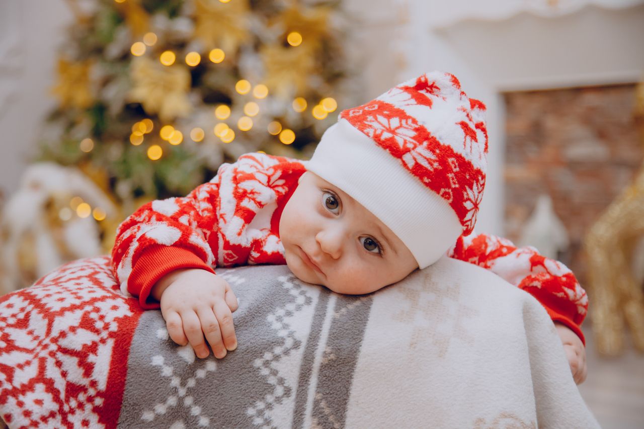 gallery-5 Charming baby in festive red attire resting beside a decorated Christmas tree indoors.