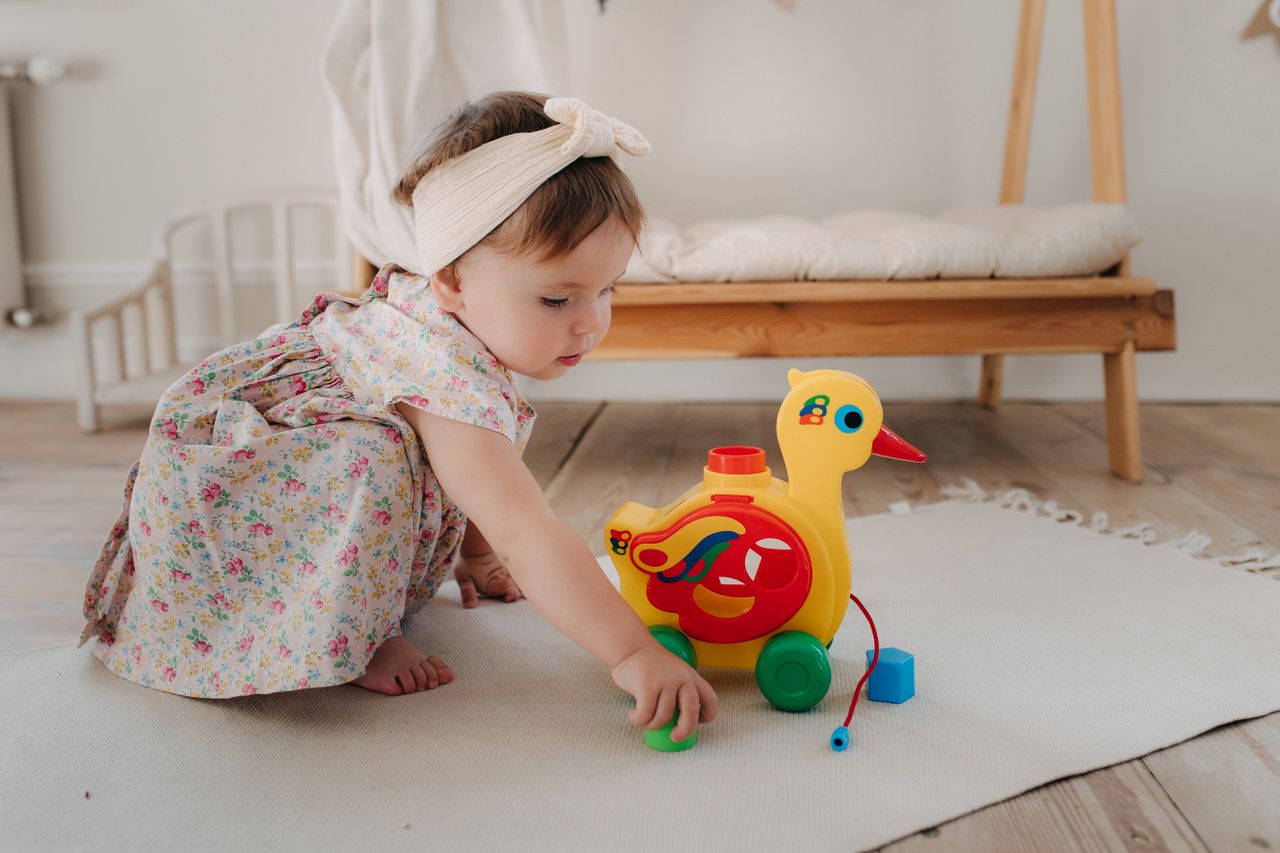 contact-img Cute baby girl playing with a colorful duck toy on a white rug in a cozy room.