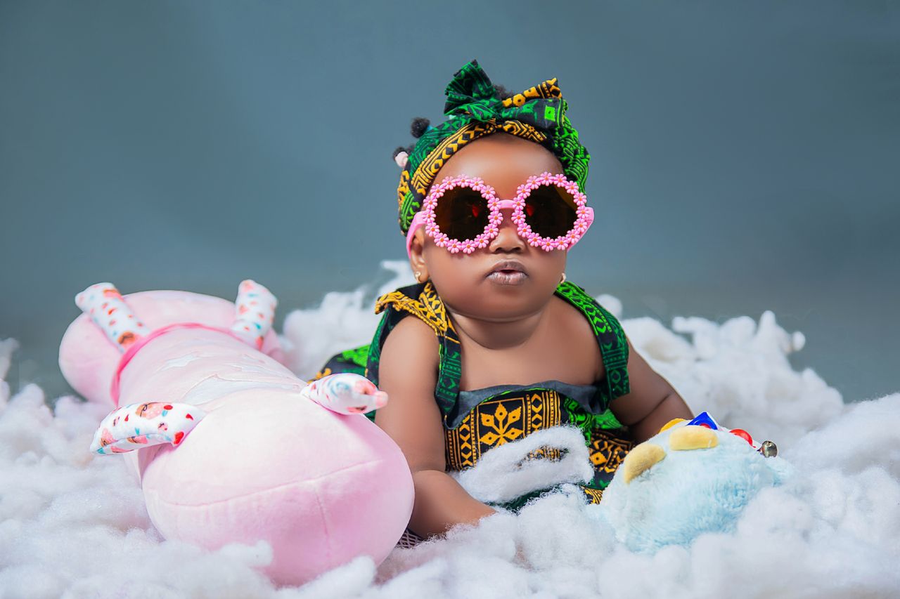 about-01 Cute baby dressed in vibrant outfit with sunglasses, surrounded by plush toys. Captivating and colorful.