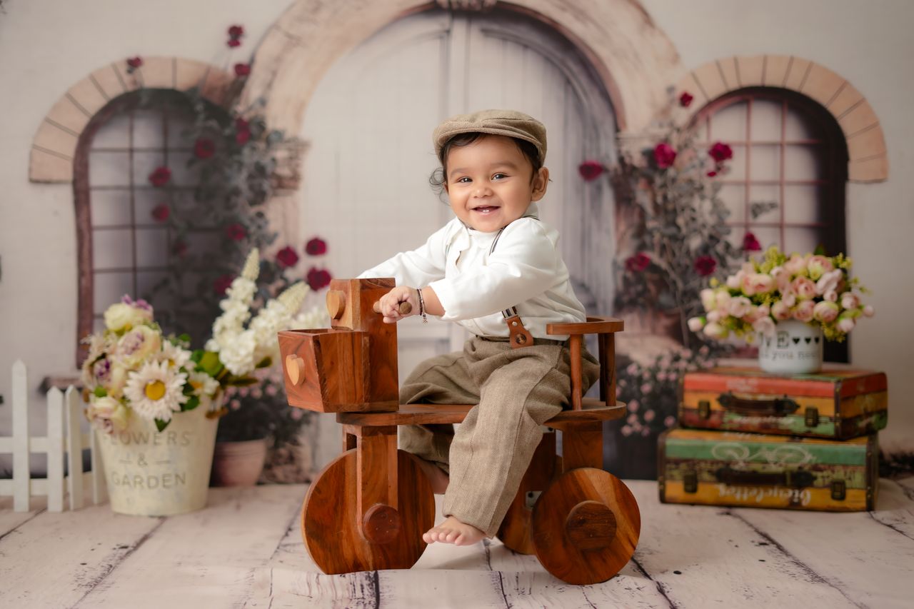 services-04 Charming toddler smiles while riding a wooden toy bike surrounded by flowers and vintage decor.